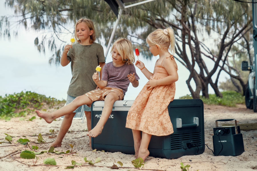 A group of 3 children holding half eaten iceblocks and sitting on top of a Dometic fridge with power pack next to it at the beach.