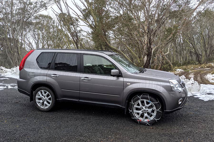 A silver-grey 4WD vehicle with snow chains fitted on the tyres. There's patches of snow on the ground and thick high country trees behind.
