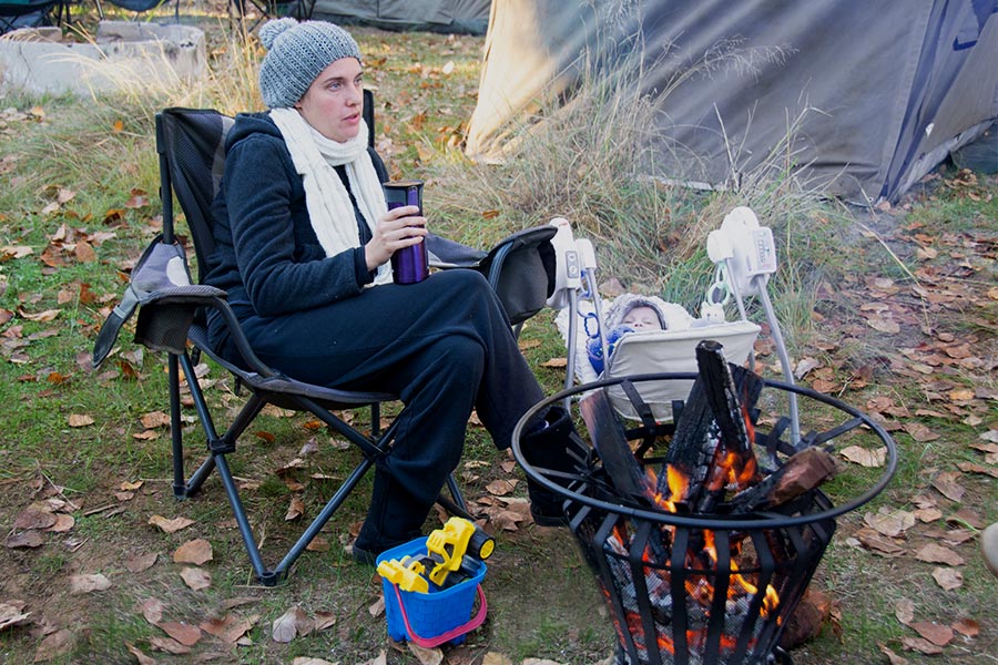 A woman sits by a campfire rugged up in scarf and beanie and holding a thermos of something hot.