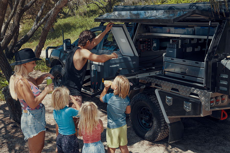 A family are gathered around a well kitted out 4WD vehicle. They are sharing a cool drink with the dad reaching in to a Dometic portable fridge.