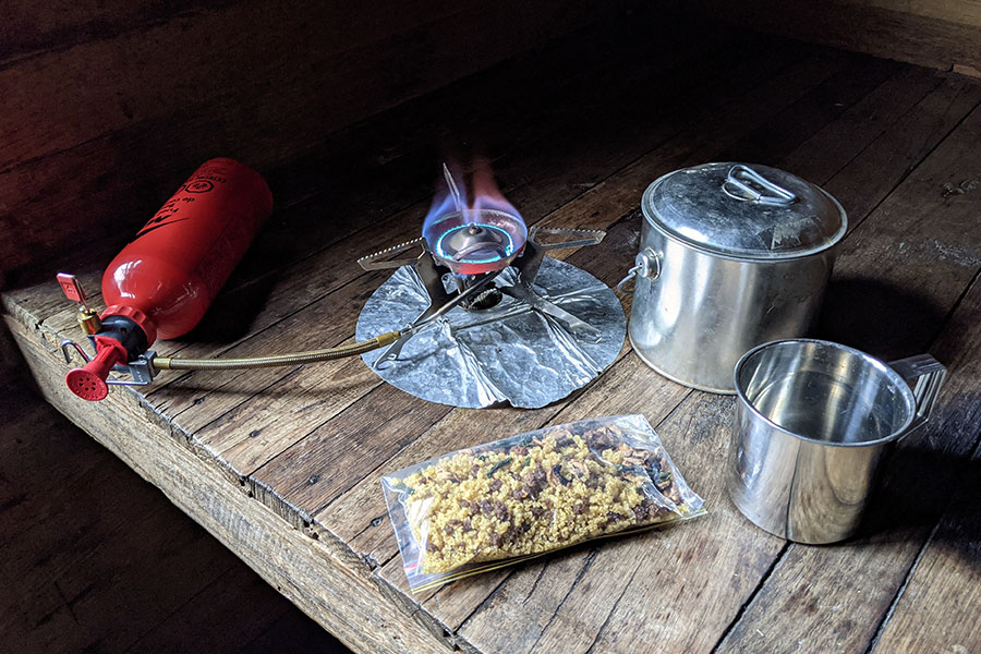 An MSR Whisperlite stove and fuel bottle is set up ready to cook dinner for a hiker. A packet of food, billy and cup sit on the bench nearby.