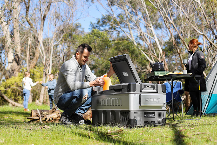 A family campsite with MyCOOLMAN dual Zone fridge in foreground. Kids play in background and a man is getting a 2 litre orange juice out of the fridge.