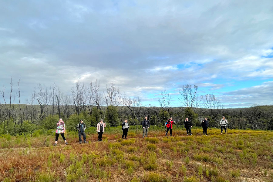 A group of nine people lined up along a walking trail and facing the camera. There's a wide spans of open land around them and regrowth from bushfires the year before.
