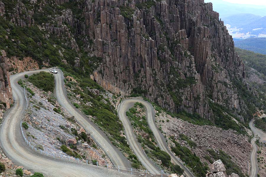 A snaking road leads down the side of a mountain with sharp cliffs and peaks behind. There's a lone white car driving down from the top.