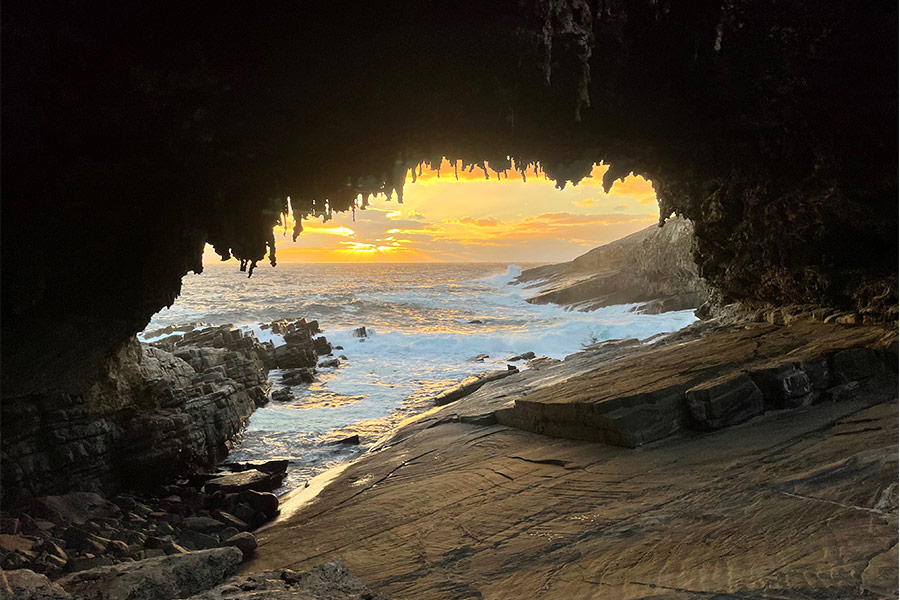 The iconic view of the sun setting through Admirals Arch. There's rock in the foreground and wild ocean crashing against the rugged coastline in the background seen through the arch.