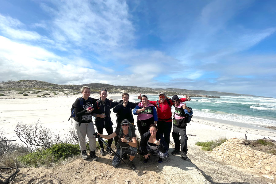A team photo of eight people at the end of a 5-day hike. Behind them is an isolated beach scene.