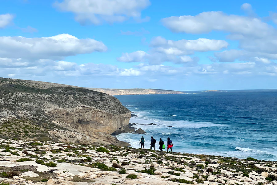 A wide shot of rugged coastline with four people walking along the rock overlooking the ocean.