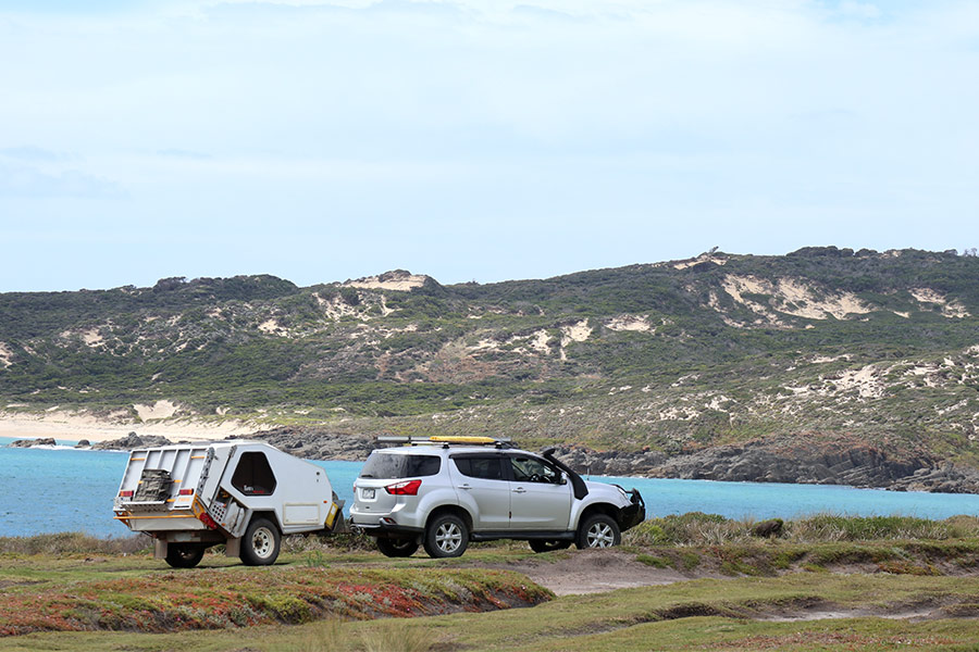 A white 4WD with snorkel is towing a camper along a Tasmanian coastal area. The blue ocean divides a foreground of green grass and a background of coastal vegetation growing amid sandy hills and dunes.