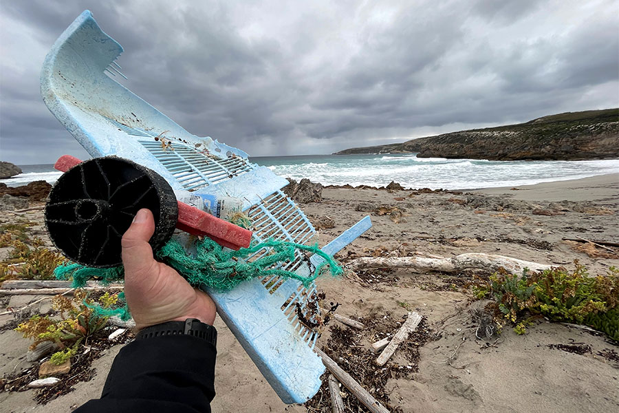 Leave No Trace - Take nothing but photographs, leave nothing but footprints. A hand holding a collection of rubbish that's been washed up onto the beach. The beach, ocean and coastline are in the background.