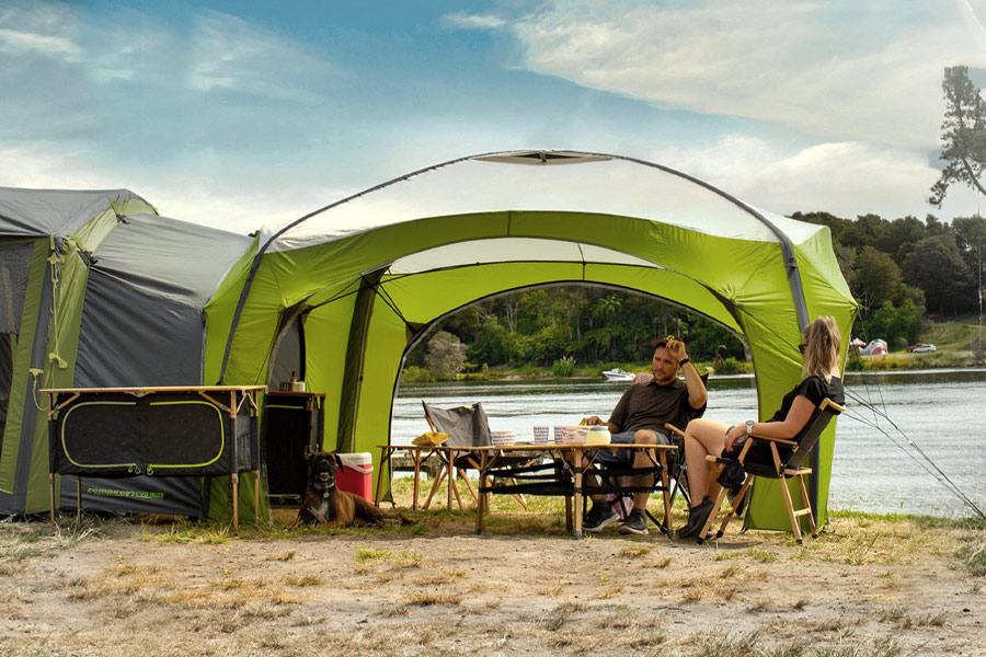 Zempire Air Tent setup with camping furniture on the bank of a river. There's a man and woman sitting back relaxing at the table.