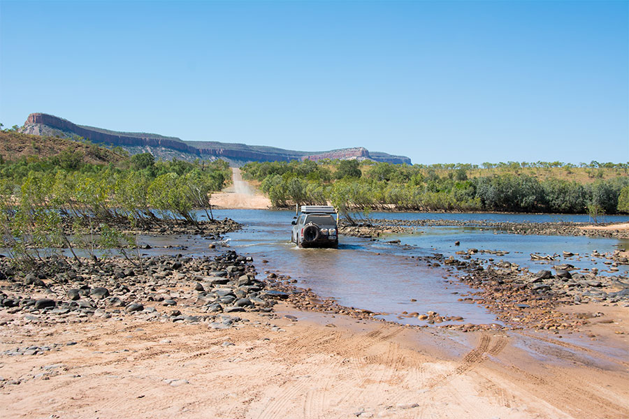 A 4WD passing through a shallow river in Outback Australia. There's a mountain range in the background.