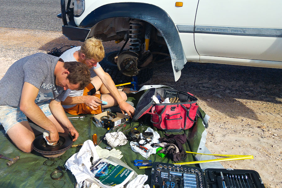Two men are sitting on a tarp with tools laid out next to them. A white 4WD is parked next to them with the wheel removed. The men are fixing it.
