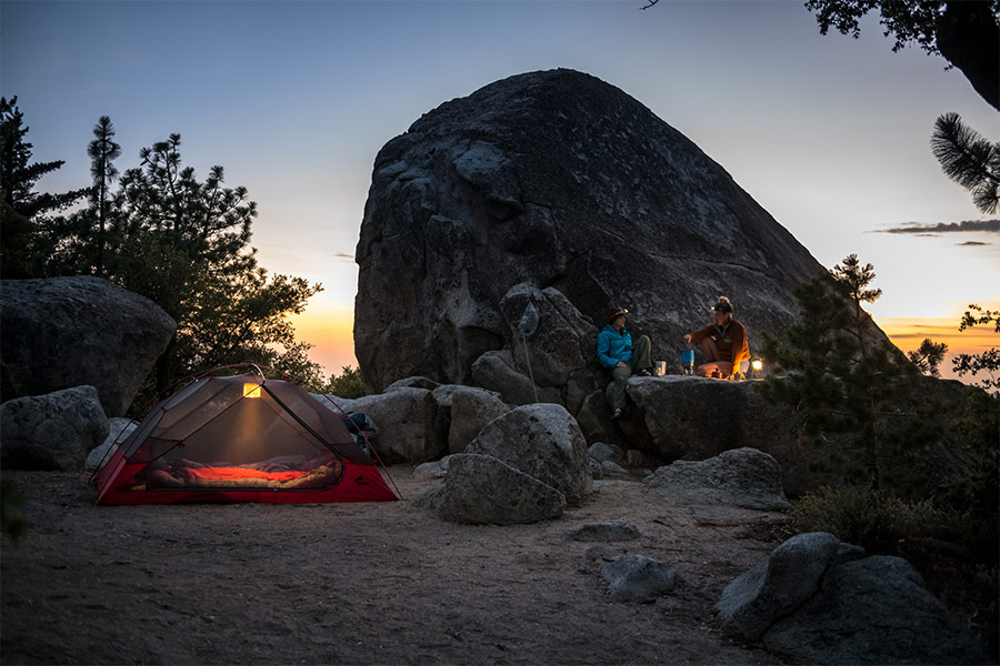 A huge boulder is the backdrop for an evening scene with 2 hikers. Their MSR hiking tent is set up to the left with a hanging lantern puttin gout a soft glow and the hikers are off to the right cooking dinner atop a rock in front of the huge boulder.