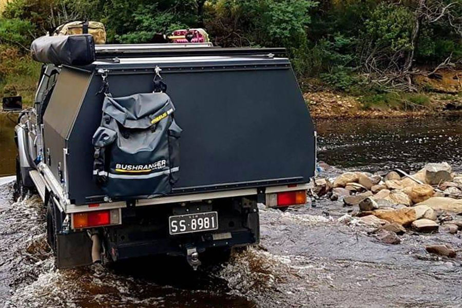 A black fully kitted 4x4 with hard canopy crosses a creek. There's a Bushranger bin bag attached to the back of the vehicle.