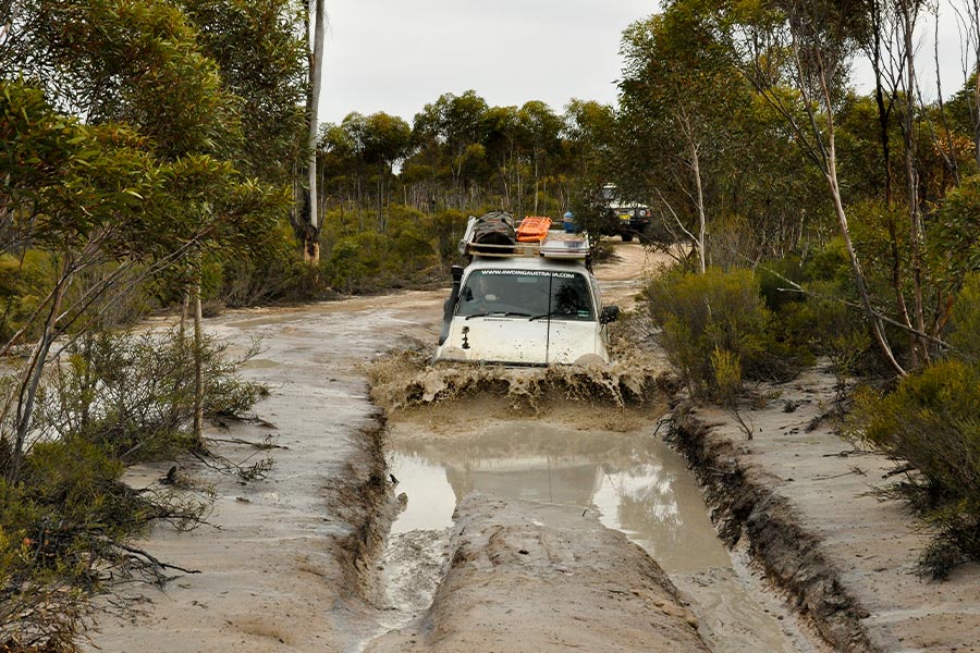 A white 4WD enters a deep muddy puddle on a track. Dirty water splashes up against the front of the car.