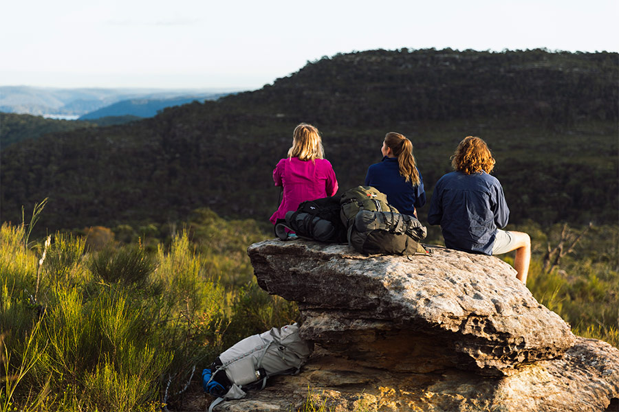 Three hikers with rucksacks lying nearby sit with their backs to the camera looking out over the view from a high vantage point.
