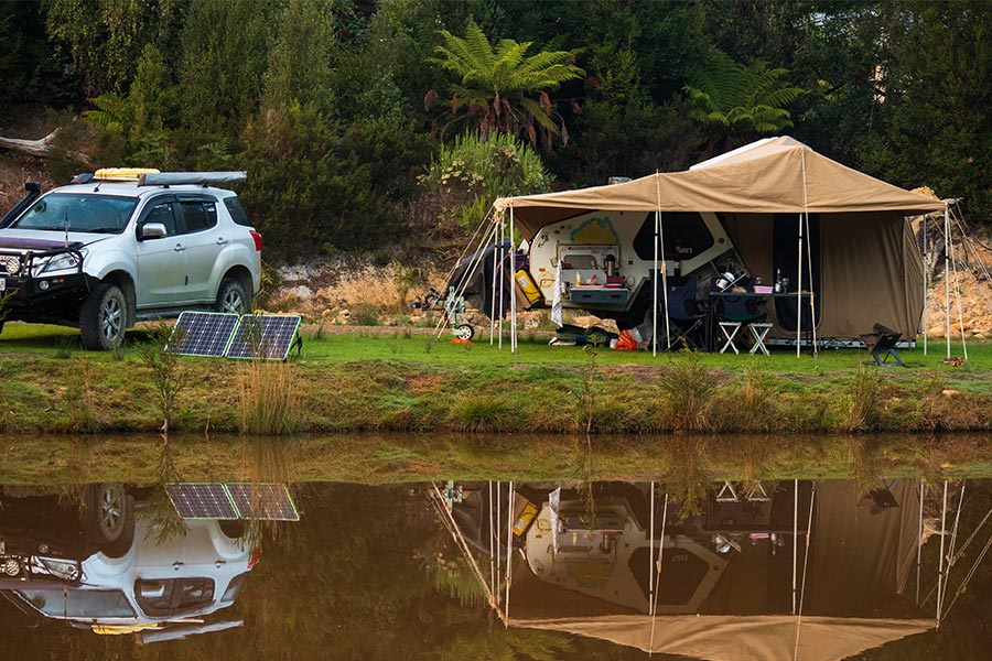 Left of Field is located at the gateway to the Mount Field National Park. A lakeside camp setup with the lake in the foreground and camper pitched on the grass behind. There's a white 4wd off to the left of frame with portable solar panels in front.