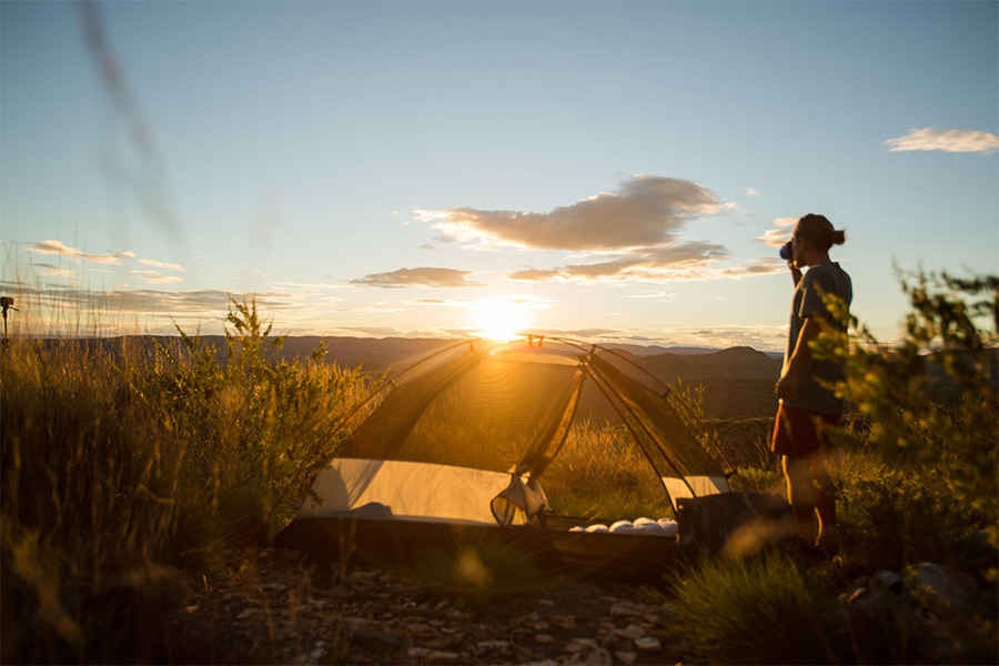 A able hiker sips a coffee while admiring a golden sunrise. He's standing to the right of frame and his hiking tent sits centred in shot without a fly. 