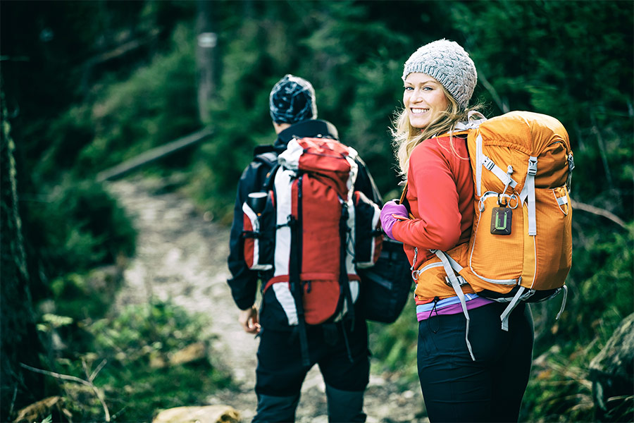 Two hikers walk along a trail with green foliage around. They are both carrying rucksacks and the female hiker has a ZOLEO Satellite Messenger attached to her orange rucksack.