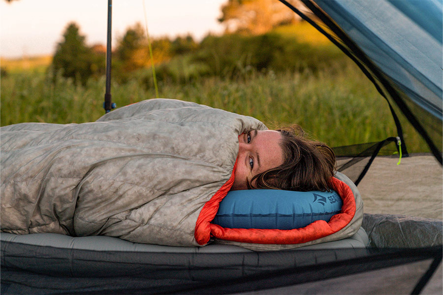 A person is tucked up inside their Sea to Summit sleeping bag with just her head peaking out. She's inside a mesh hiking tent and lying on a mat with an inflatable pillow. We can see morning light a scrub in the background.
