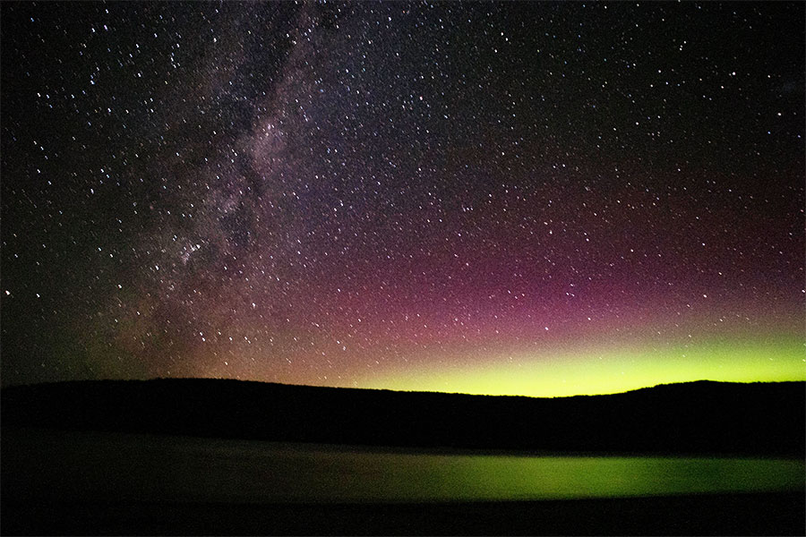 The Southern Lights against a starry sky. Hues of green, yellow and magenta mark a striking silouette of land on the horizon.