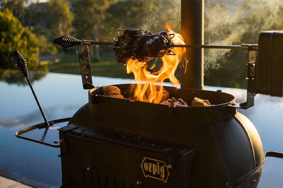 Close up image of the Big Pig cooker by Ozpig set up with the spit rotisserie accessory. There's a fire going and a large cut of meat cooking in the flames suspended above by the spit accessory. Trees and the glow of late afternoon sun are in the background. 