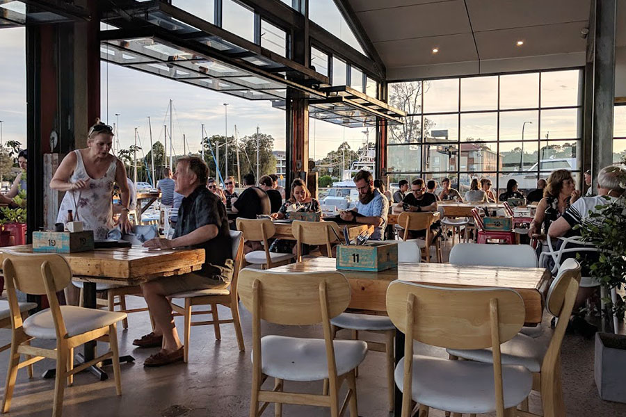 A cafe scene with tables, chairs and diners. There's glass windows all around with light streaming inside and bifold doors wide open for alfresco seating.