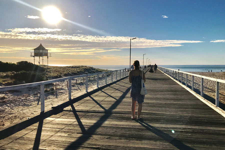 The sun is setting and casting long shadows across a jetty as a woman with her back to the camera walks away down the jetty toward the end.