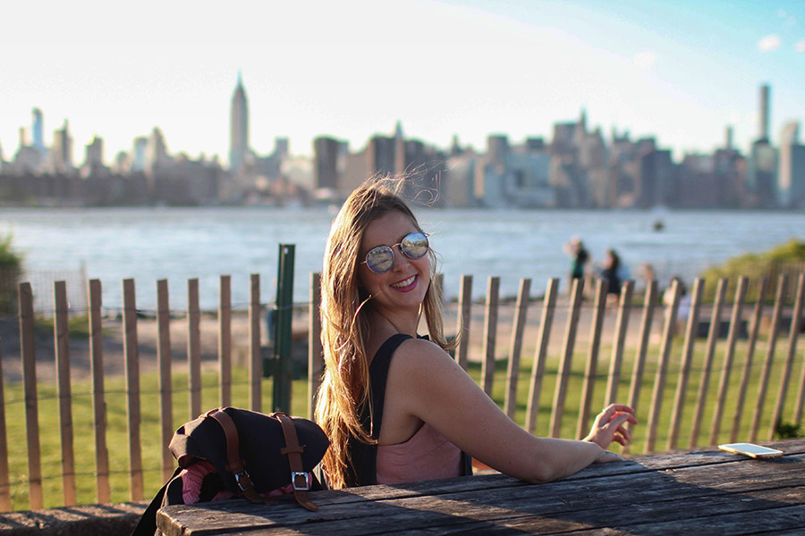A woman is sitting at a park bench looking over her shoulder and smiling at the camera. She is wearing reflective sunglasses and there is water behind her with the tall buildings of a cityscape in the background.