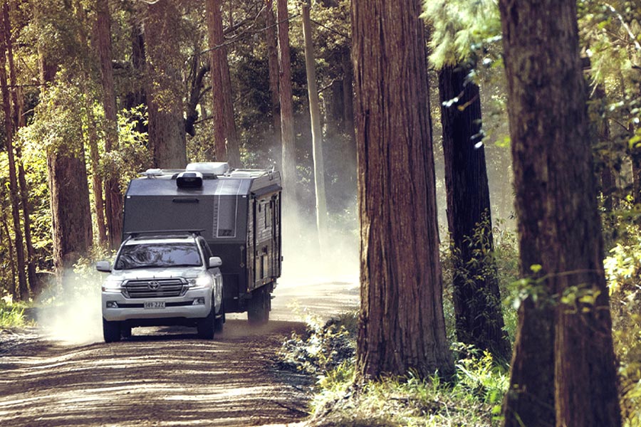 A white 4WD is towing a large black caravan along a dusty forest track. There are trees either side and a cloud of dust behind.