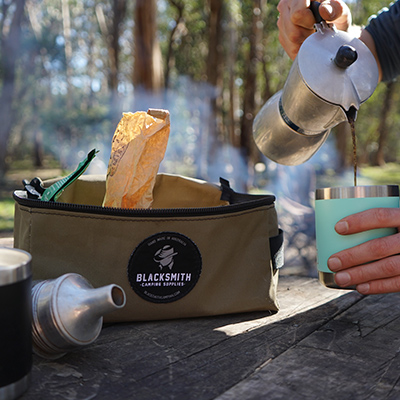 Close-up of a morning brew setup in the outdoors. A hand pours coffee from a stovetop percolator into a travel mug and an open Coffee Kit Bag from Blacksmith Camping Supplies, shows a few coffee making accessories. It all sits on top of a rustic wooden table and there are blurred trees in the background.
