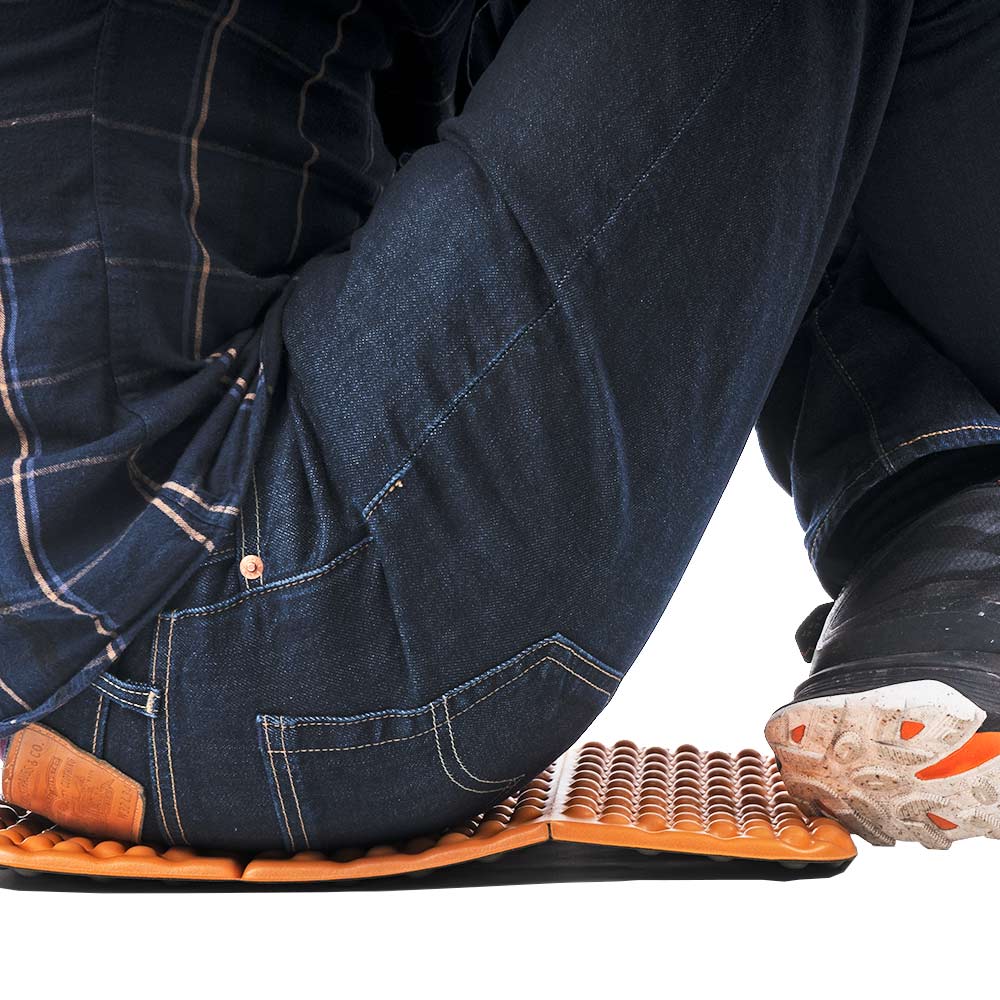 A man wearing dark denim jeans and a dark checked shirt sits crosslegged on the orange Sit Pad Flex.
