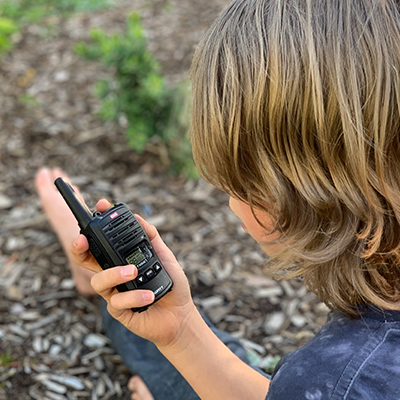 A close-up shot looking over a child's shoulder at their raised hand holding a GME UHF CB Handheld Radio. There's a burred background of bark chips and a small bush.