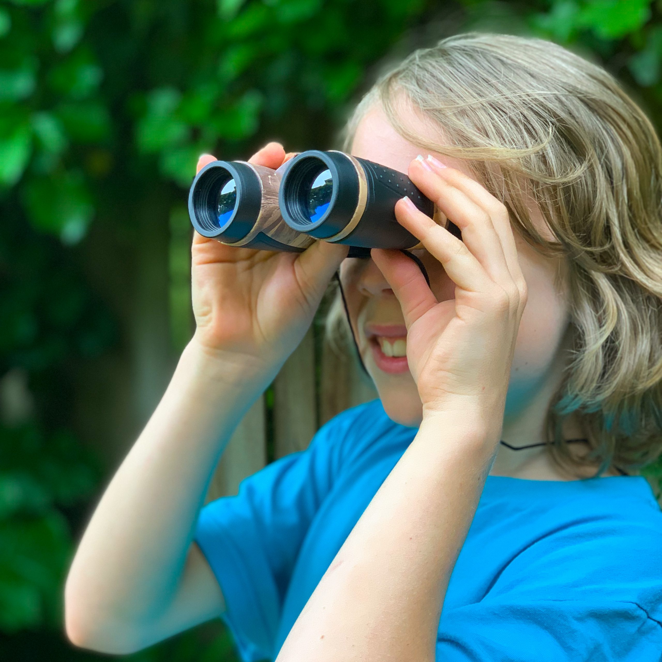 A close-up of a young boy wearing a blue t-shirt and holding a set of kids binoculars to his eyes. He's smiling at what he sees and there is lush Gree foliage in the background.
