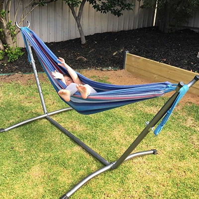 The Anywhere Hammock Double with Frame is set up on a lawn with garden bed in the background. A young child is wrapped up inside the hammock, laughing with legs and arms poking out.