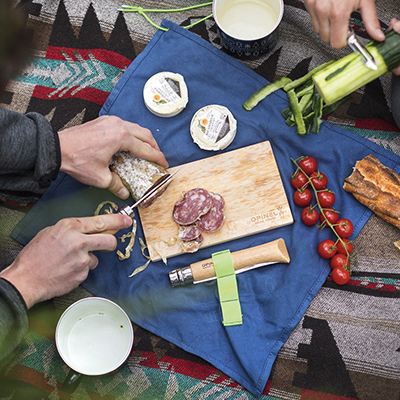 A picnic spread using the Opinel Nomad Cooking Kit. A male's hands are cutting salami on the cutting board while another set of hands is using the peeler to take the skin off a cucumber. There's also a baguette to the right of frame and some cherry tomatoes. All of it is spread out the the blue cloth that comes with the kit.