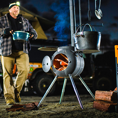 It's nighttime as a man wearing a headlamp carries a big pot to an Ozpig Cooker Heater. There's a fire going inside the cooker and other pots and utensils around.
