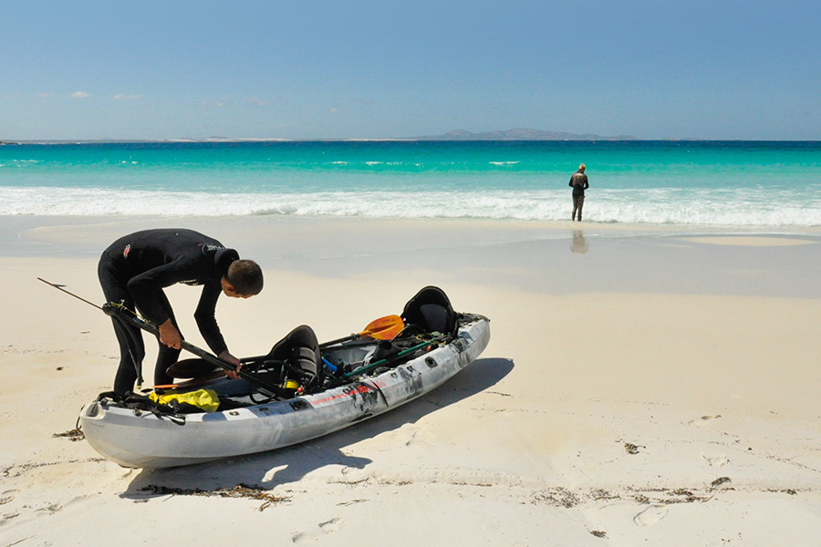 A man leans over a kayak to prepare his gear while another person is off in the far distance standing by the waters edge. They're on a beach with white sand and turquoise water.