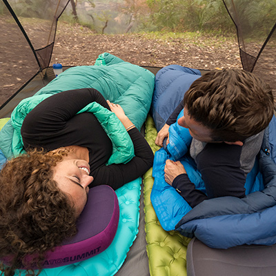 A male and female lie inside sleeping bags in a mesh tent. Their sleeping bags are aqua and blue, and they have blue and green Sea to Summit sleeping mats and air-filled pillows. There are ferns and green foliage outside in the background.