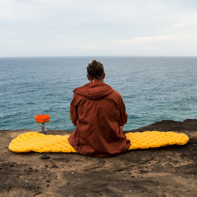 A female hiker wearing a weather jacket, sits on her yellow Ultralight sleeping mat. She has her back to the camera and is looking out to sea from a rock platform.