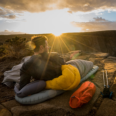 A male hiker wakes up to a bright morning sun rising just above some rocky cliffs by the ocean. He's camped on the cliffs with a Sea to Summit hiking mat, pillow and sleeping bag.