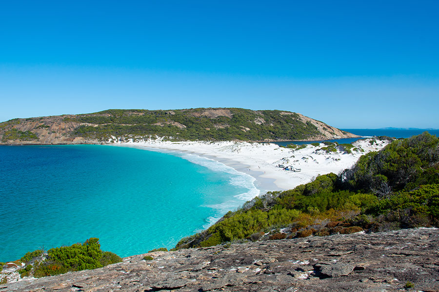 An idyllic bay with turquoise water and a white sandy beach. There's a rocky platform in the foreground, blue sky above and a small island on the horizon.
