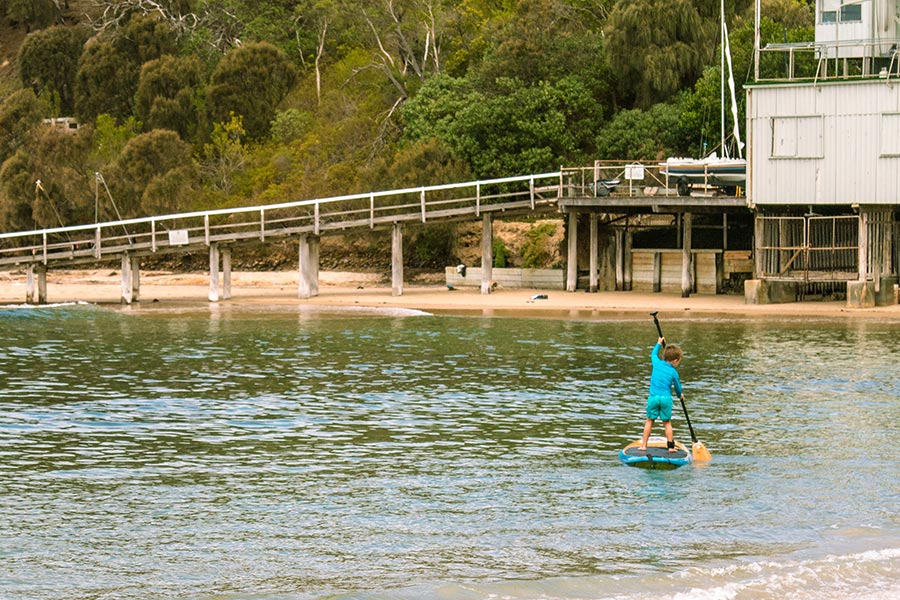 A young boy practices his SUP technique in shallow water at Pt Leo. There's a jetty in the background running from the old yacht club, and a thickly treed hill rising out of frame behind.