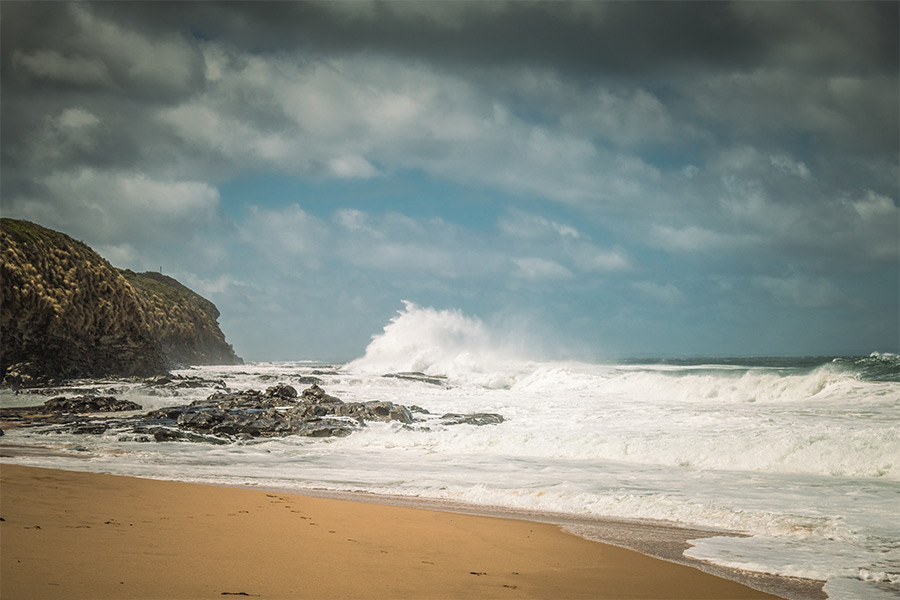 A wild surf break with stormy dark clouds overhead and wet sand in the foreground. The rocky cliffs of the coastline jut out to the left of frame.