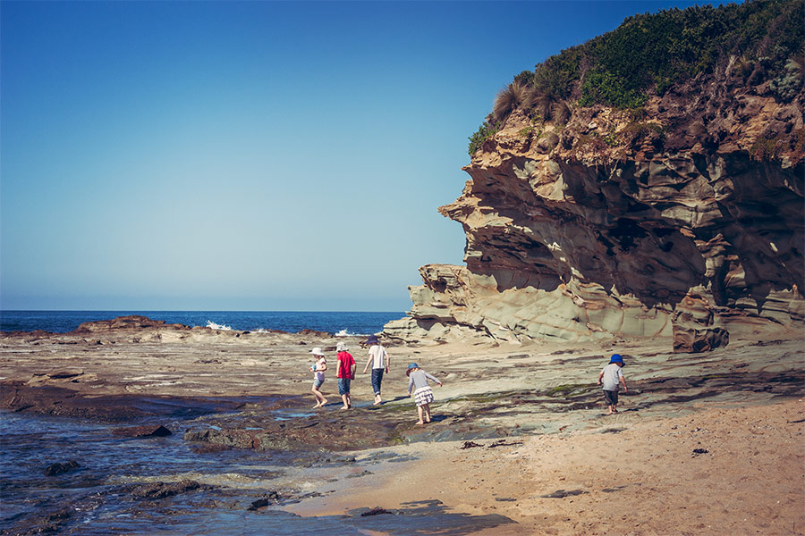 A jaggard cliff face is to the right of frame which gives way to rock pools and shallow water at the photographers level. There is a group of 5 small children exploring the rock pools and it's a clear blue sky day.