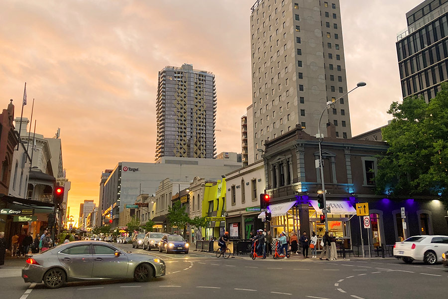 Picture of an Adelaide City street at dusk with golden tones from the setting sun. There are buildings, traffic lights and cars with headlights all around.