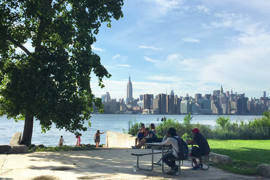 The cityscape of New York is in the background with the river stretching across the frame in front. In the foreground is a picnic table with people sat and a large tree to the left.