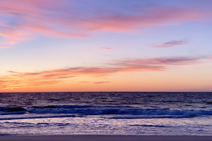 The pinky-purple hues of sunset stretch across the sky and ocean as gentle waves break close to the shore.