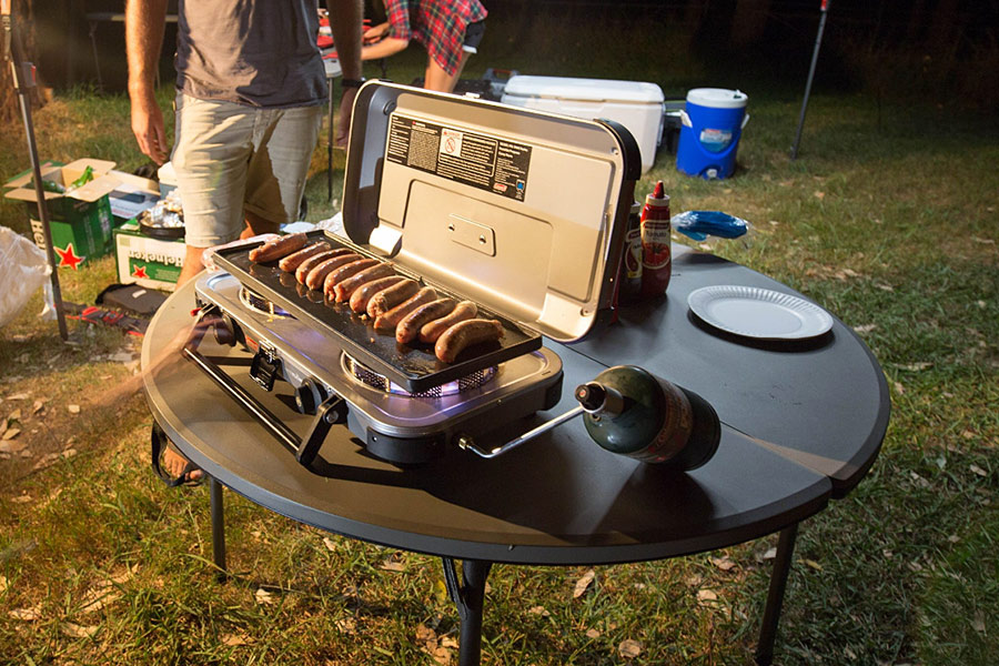 A camp stove is resting on a table at a campsite, with sausages cooking on top. Nearby is a man dressed in a grey t-shirt and pale shorts, as well as a woman in a red and navy checked shirt. It is night time.