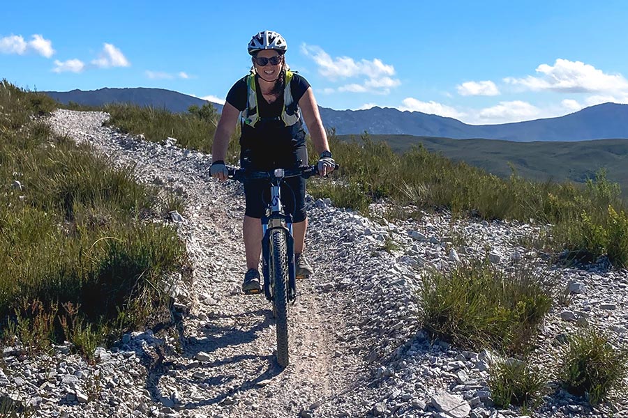 A female MTB rider faces directly towards the camera, mounted on her bike. She's on a downhill gravel track with a little low shrubbery either side and mountains in the background.